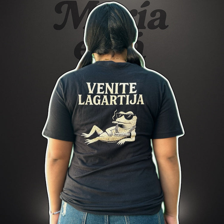 Person wearing a black t-shirt with 'Venite Lagartija' text and graphic on the back, standing against a dark background.