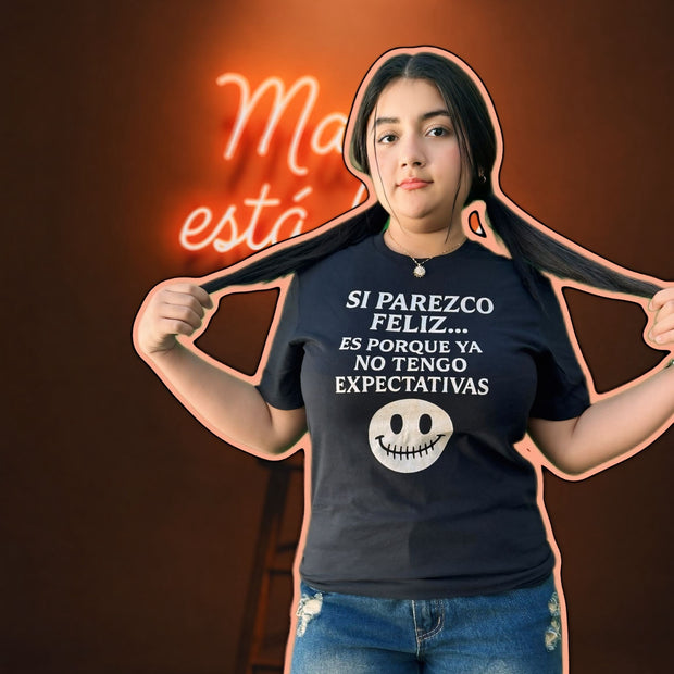 Person wearing a dark blue t-shirt with text and a smiley face graphic, standing against a dark background with a neon sign.